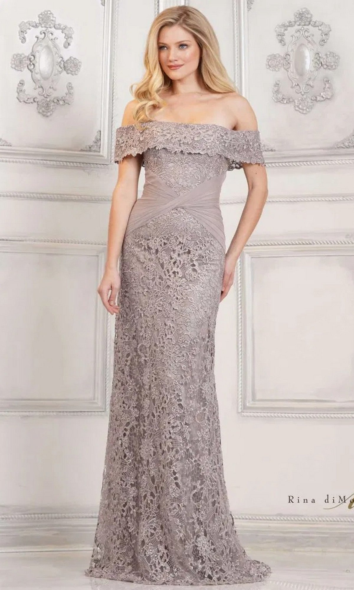 A woman in the Long Formal Dress: Rina Di Montella RD3129, an off-the-shoulder lace gown in silver, stands gracefully in an ornate white room.
